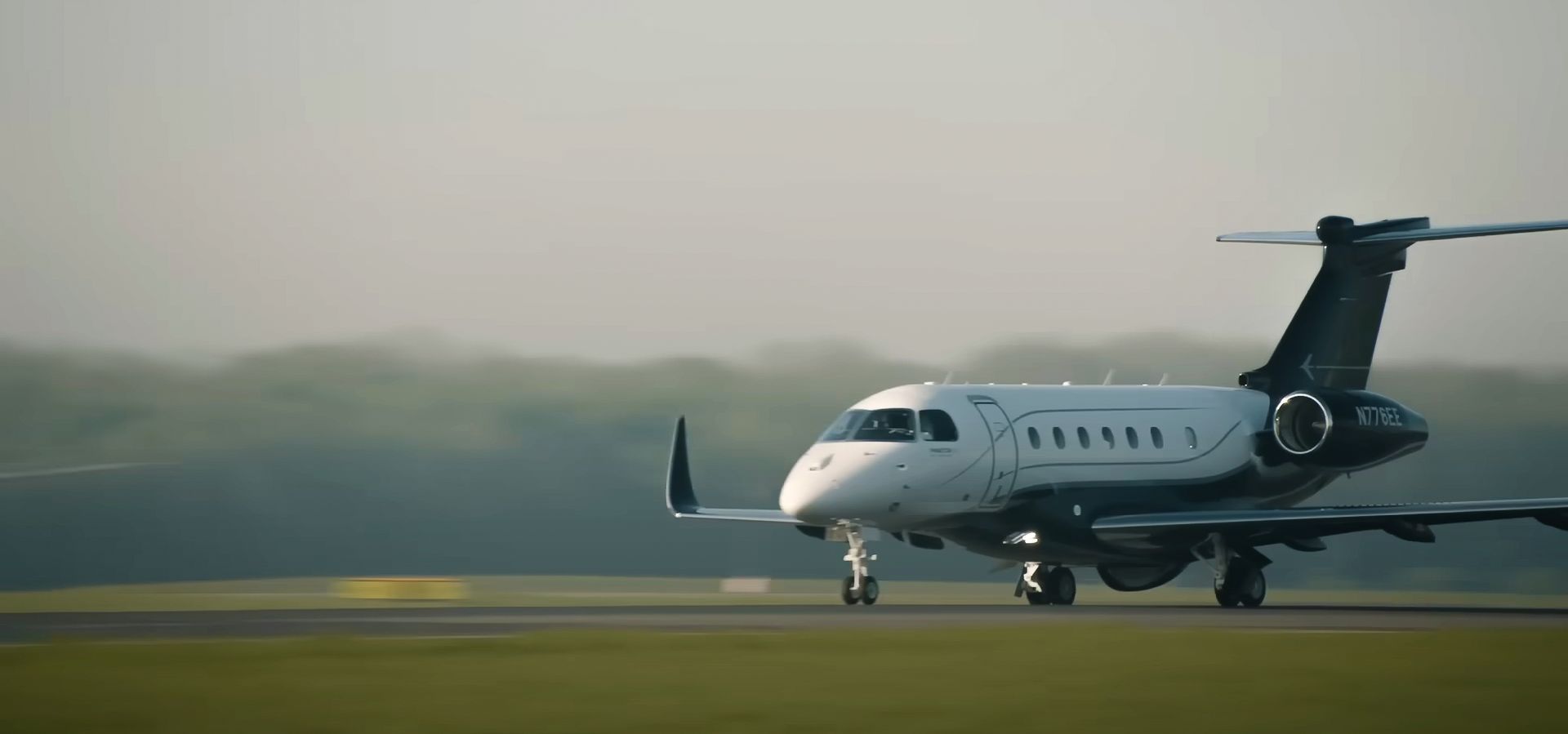 A private jet on a runway, in front of a green field and a blurry tree line under a hazy sky.