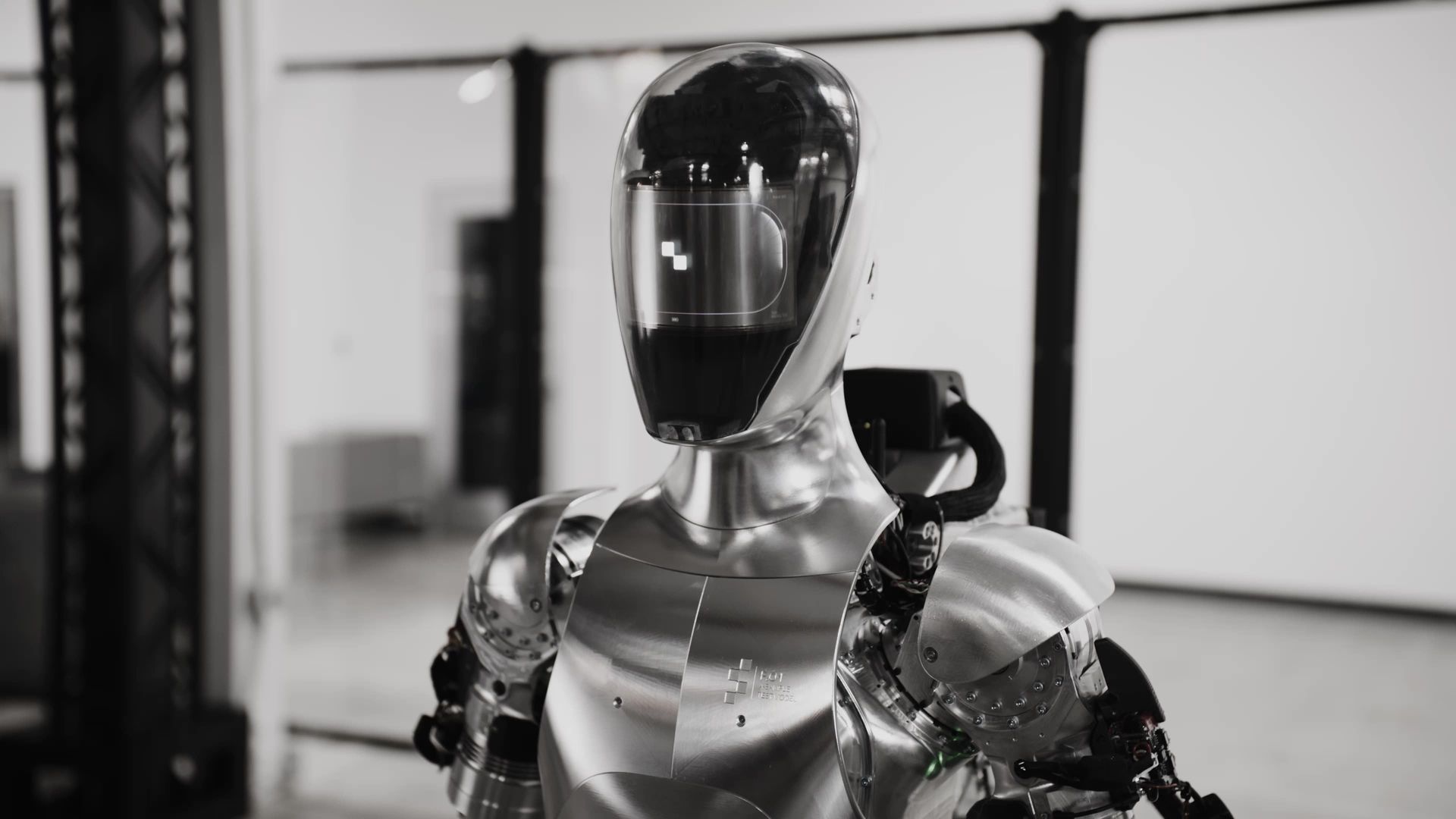 A humanoid robot with a reflective silver body stands indoors with arms at its sides, looking forward.