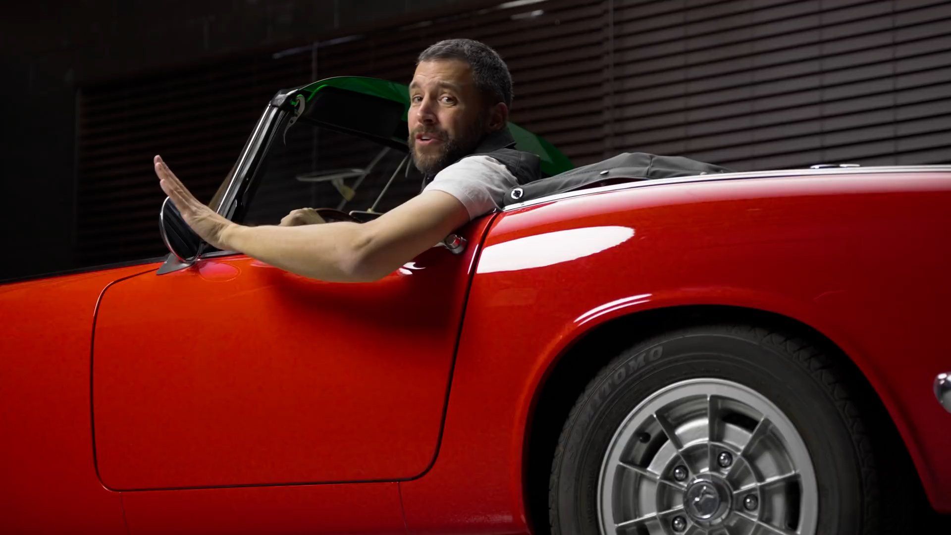 Man in red convertible waves, appearing surprised. Dark garage setting.