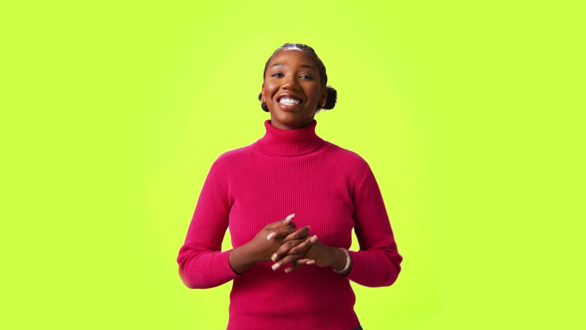 Woman in a pink turtleneck smiles, hands clasped, against a bright green background.