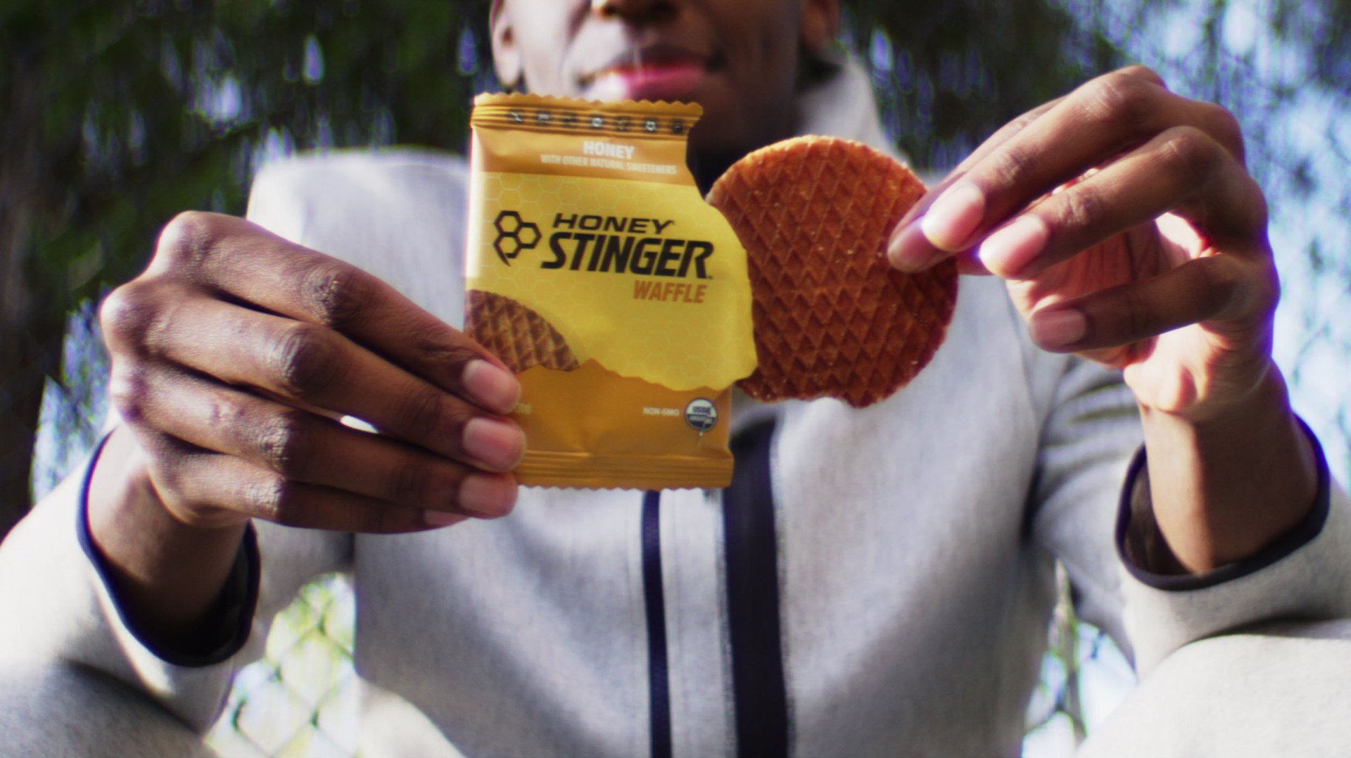 Person holding a Honey Stinger waffle, opened wrapper visible. Outdoors, bright.