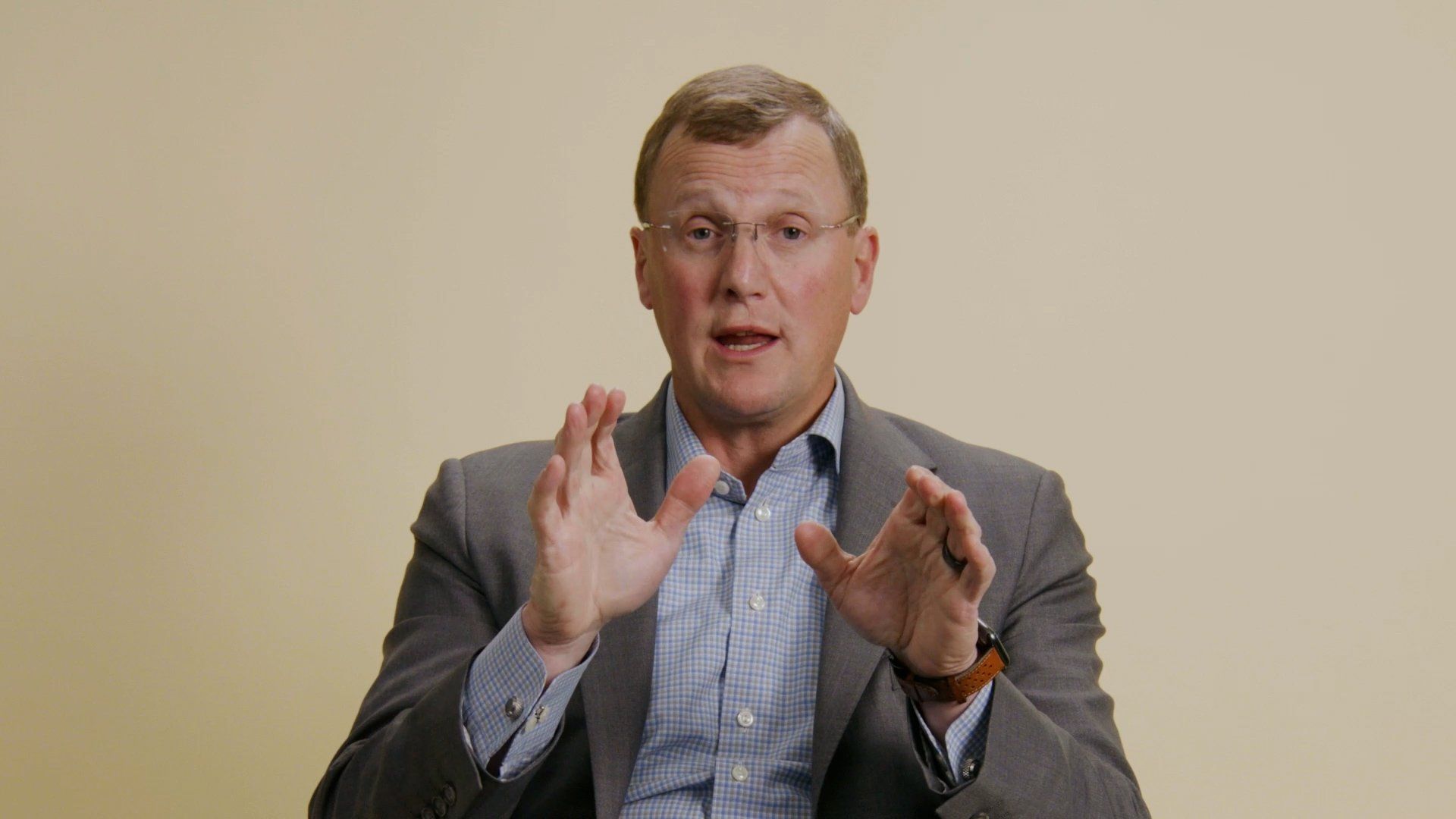 Man in a suit gestures with hands, speaking, against a light yellow background.