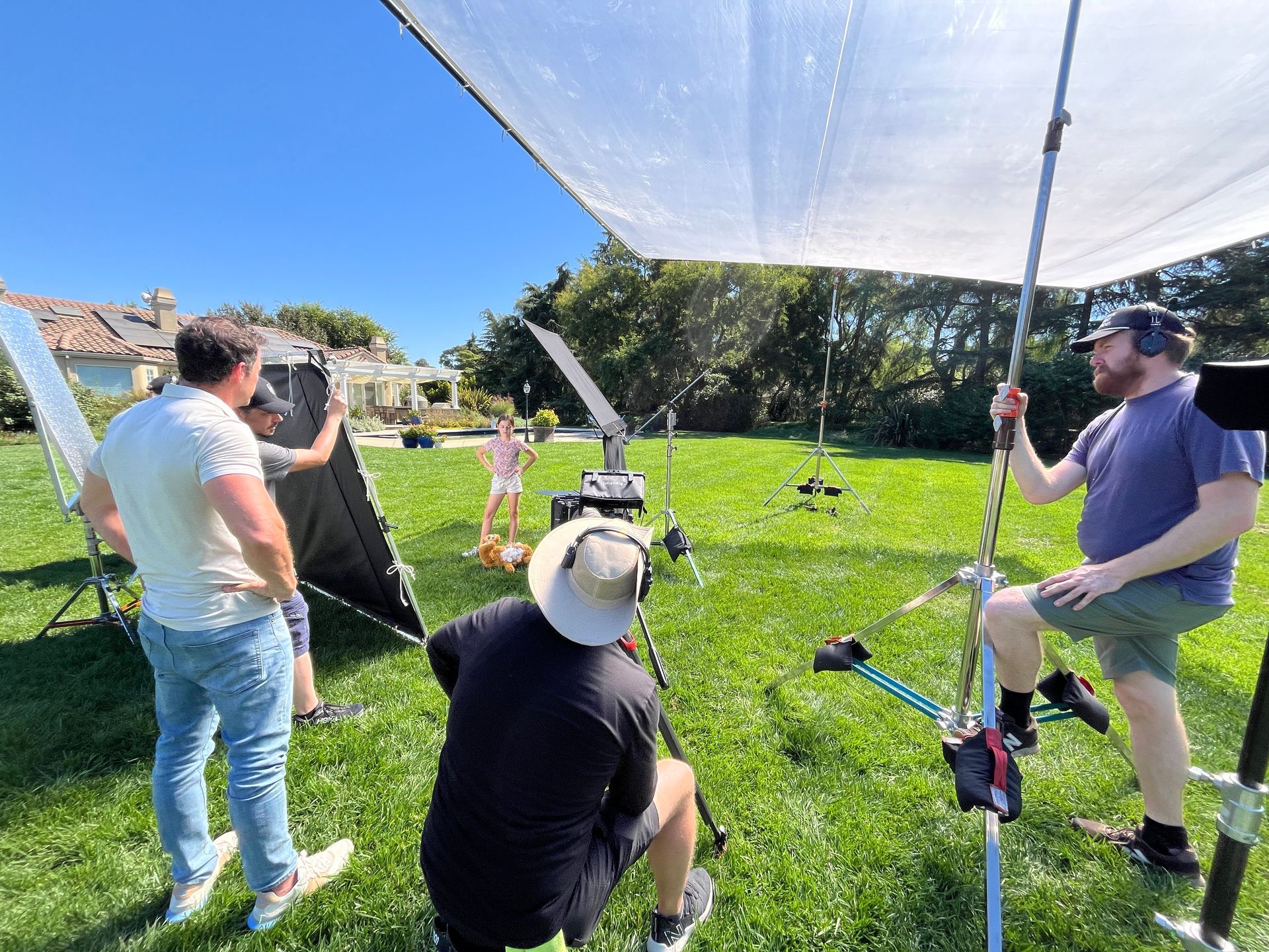 Film crew filming outdoors in a sunny park, with a white reflector and camera setup on the grass.