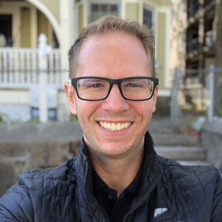 Man with glasses smiling outside, wearing a black jacket; blurred background of a house.