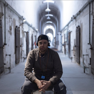 Man seated in a prison hallway, wearing headphones, looking forward.