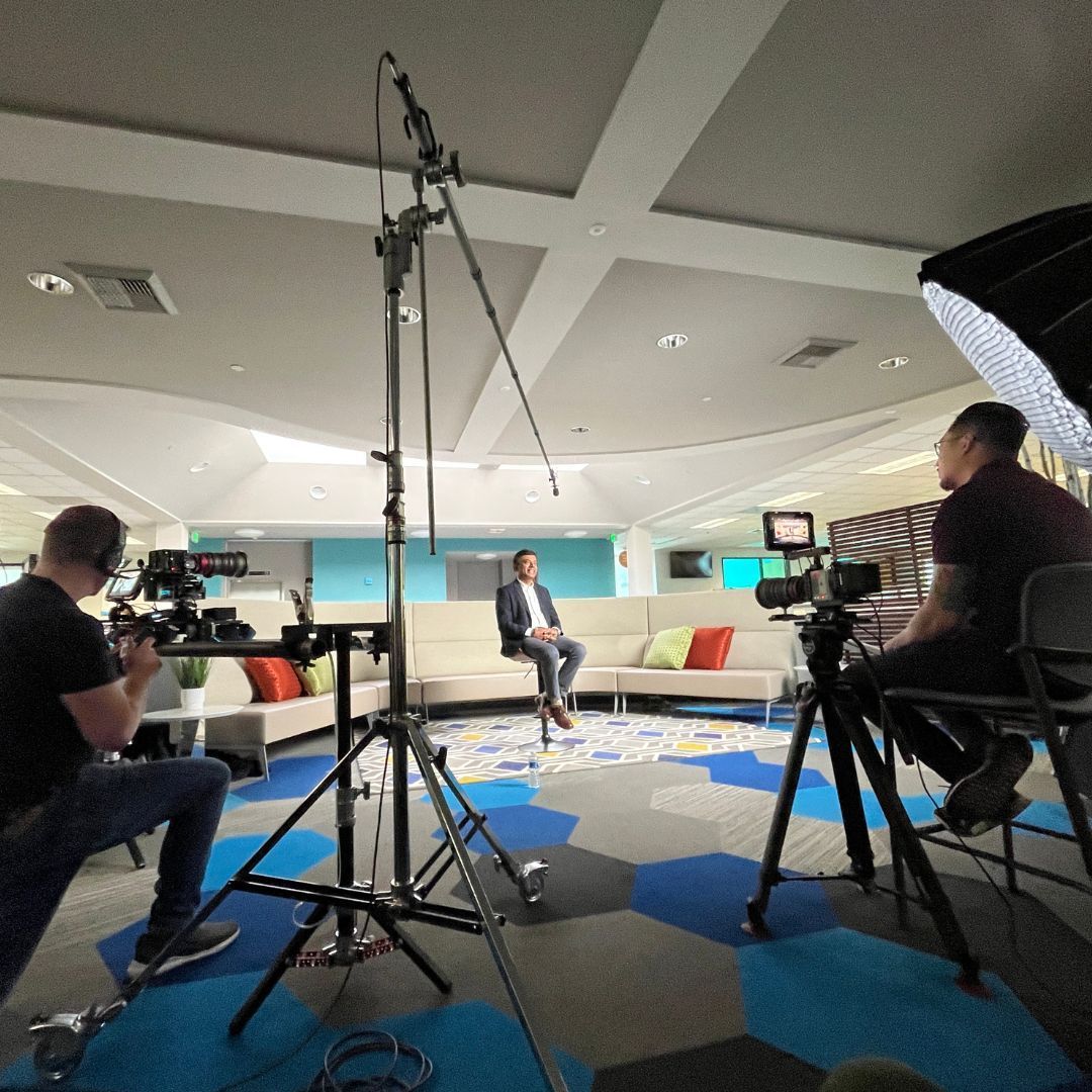 Filming an interview: A man sits on a stool, surrounded by cameras and crew in a brightly lit room.