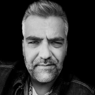 Man with graying hair and beard looks at the camera with a neutral expression, dark background.