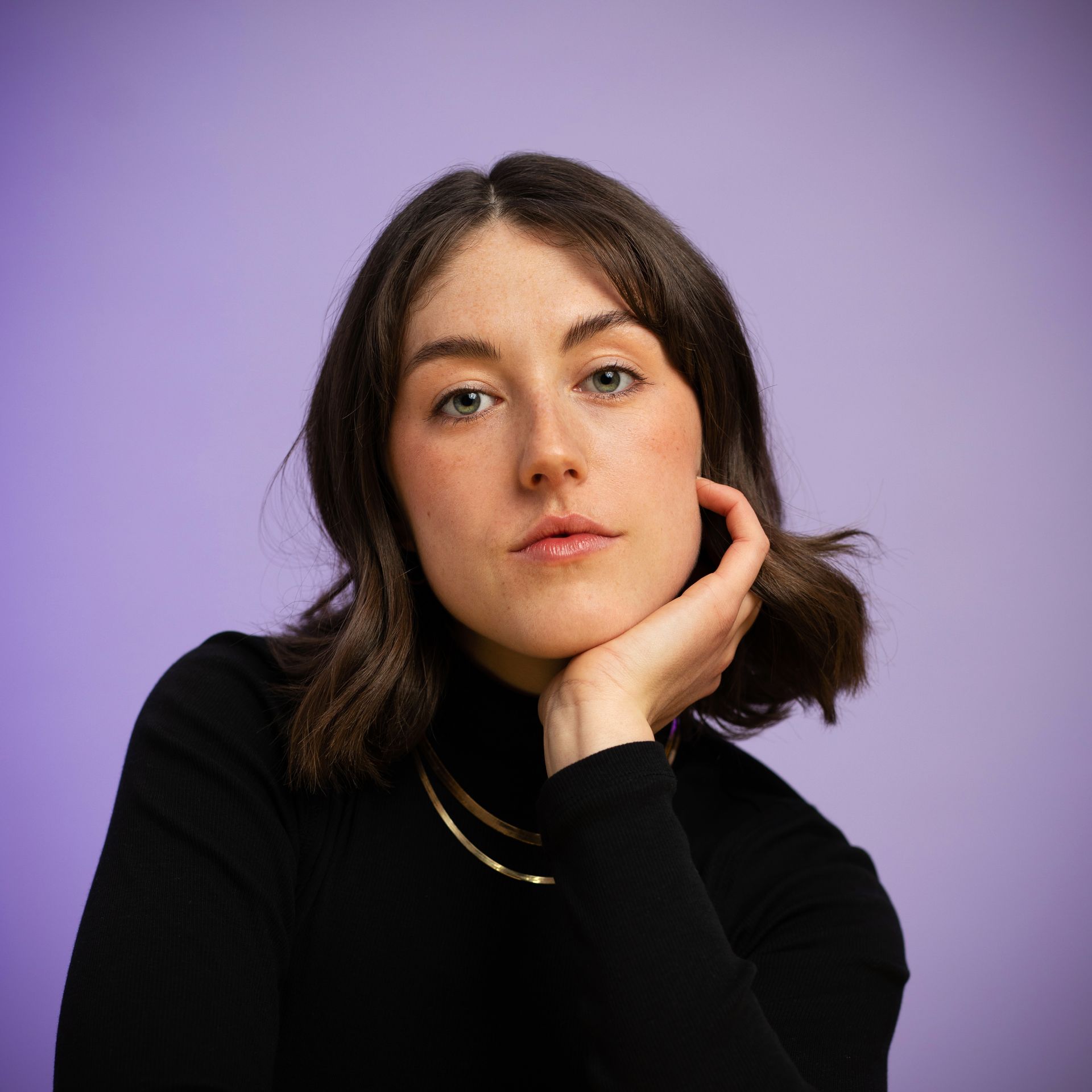 Woman with dark hair, resting chin on hand, wearing a black turtleneck and gold necklace against a purple background.
