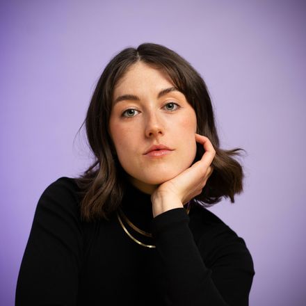 Woman with dark hair in black turtleneck, hand on chin, against a purple background.