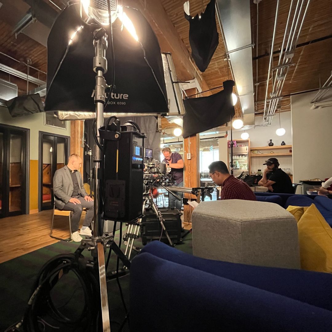 Filming a man seated for an interview. Studio setting with lights, crew, and lounge furniture.
