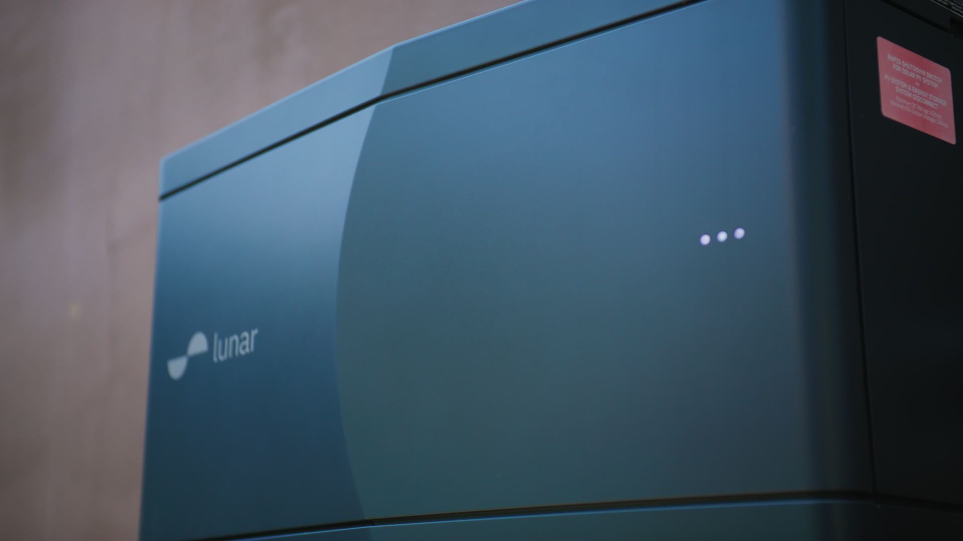 Dark teal Lunar home energy storage unit with three illuminated dots, against a neutral wall.