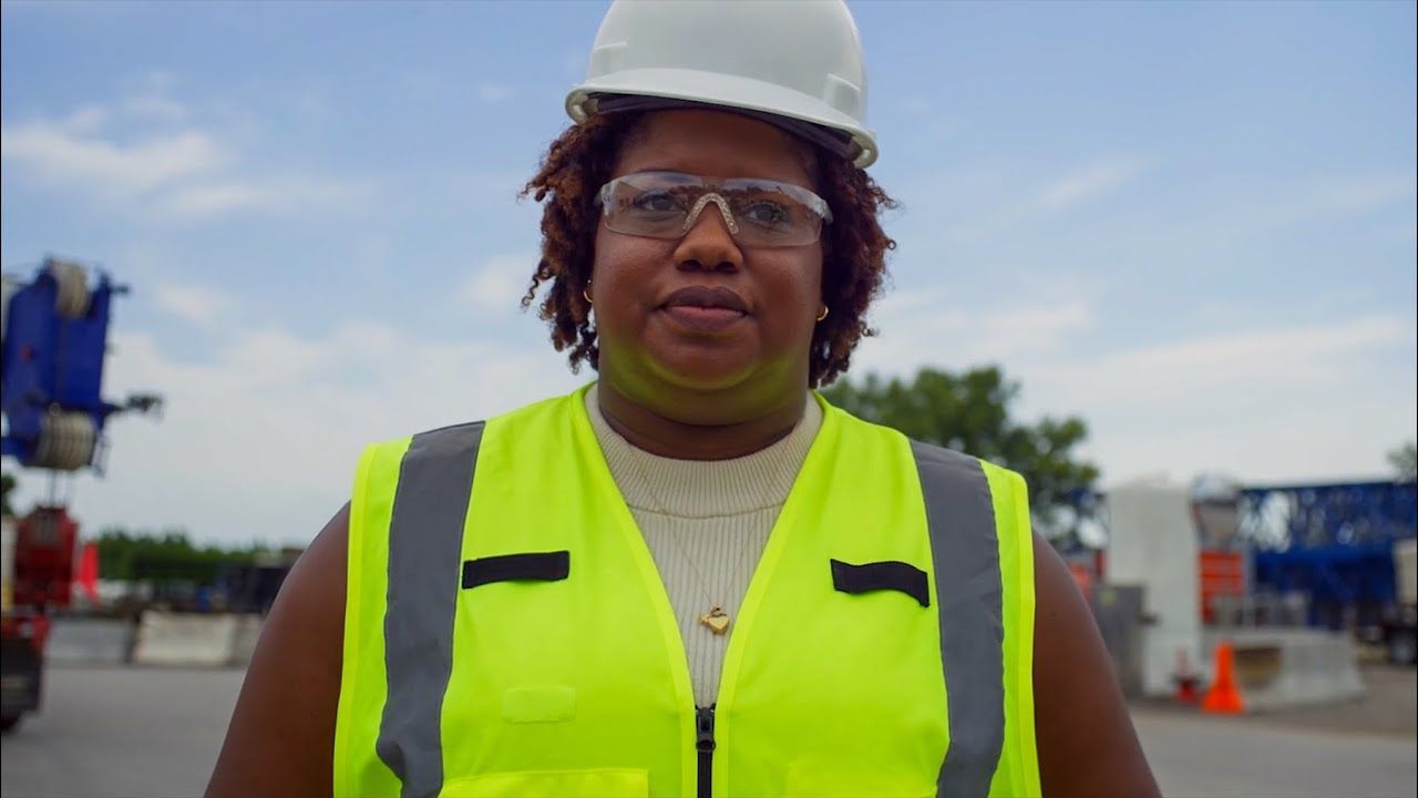 Woman in hard hat and safety vest smiles on construction site.