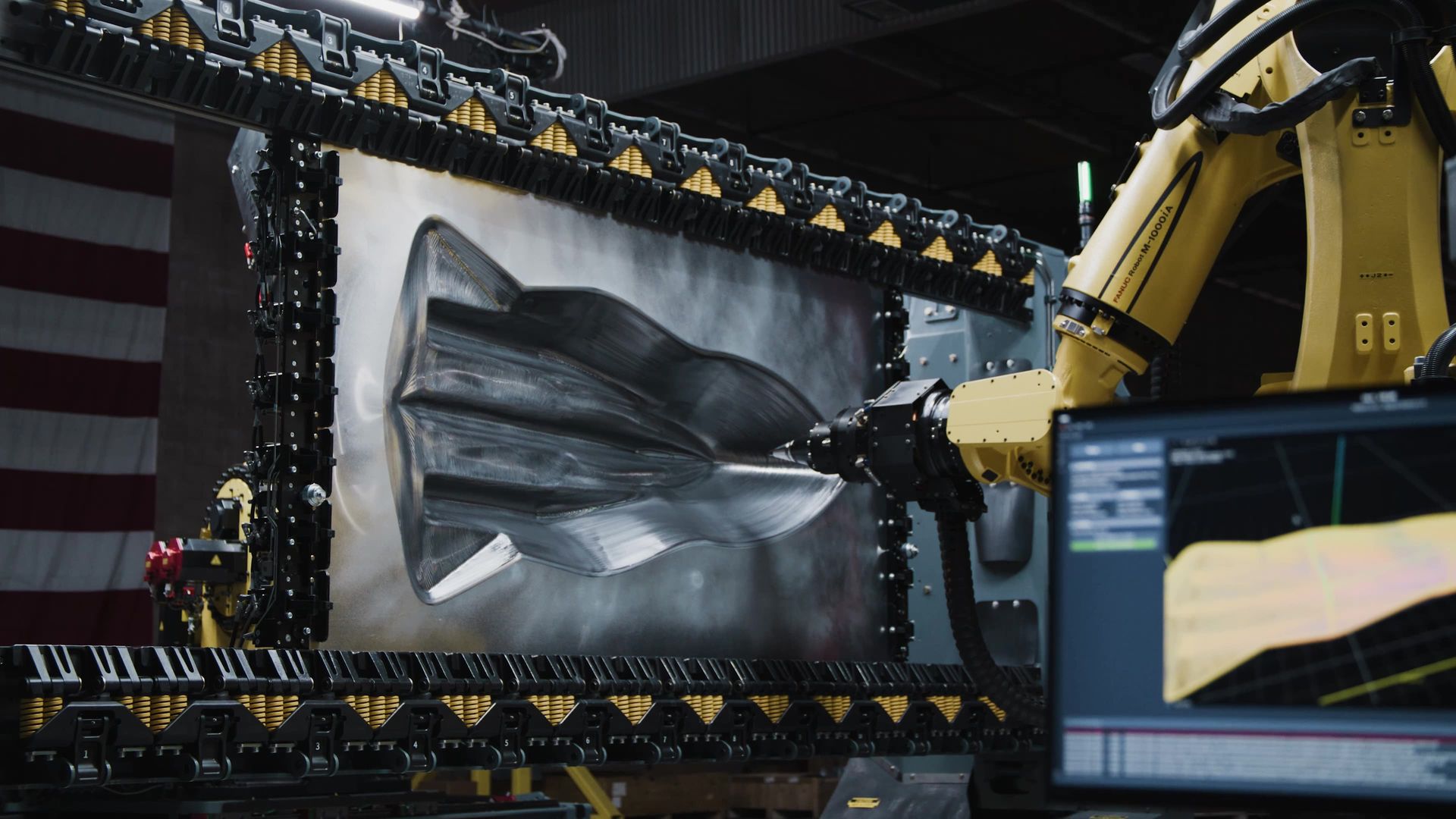 A robotic arm molds a metal object inside a large machine; American flag in the background.