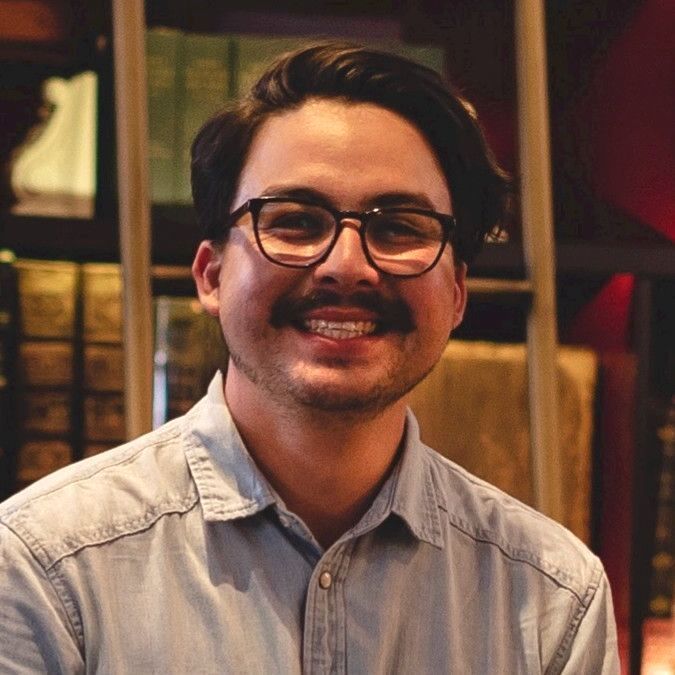 Man with glasses and mustache smiling in front of a bookshelf, wearing a light blue button-down shirt.