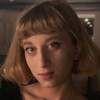 Woman with short, light-brown hair and bangs, wearing eyeliner, looking toward the camera.