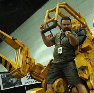 Man in olive shirt pointing while sitting in a yellow power loader, likely at a convention.