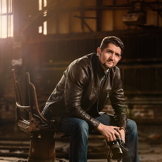 Man in leather jacket holding a camera, seated in a rusted chair inside a dimly lit industrial space.