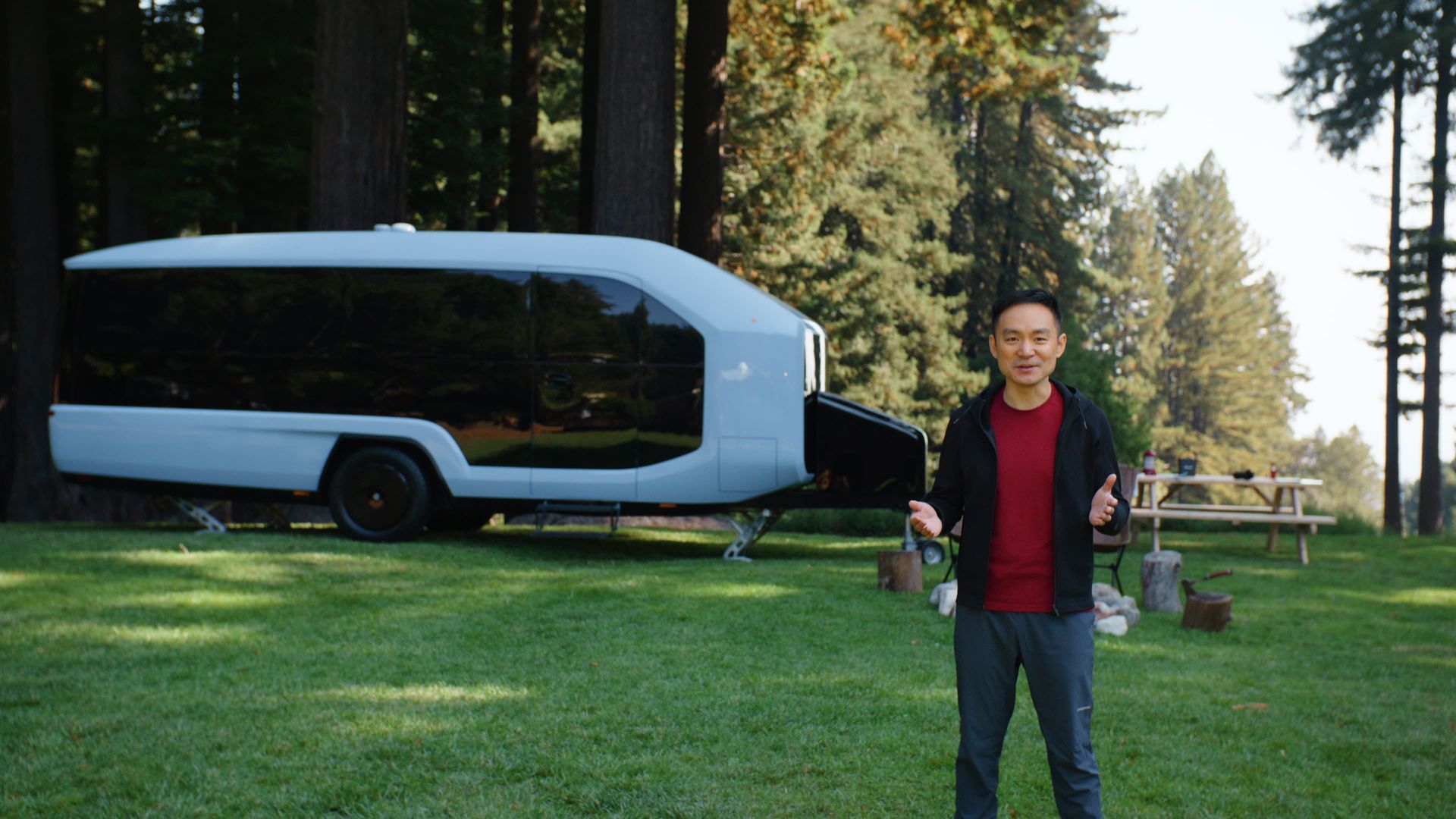 Man standing in front of light blue camper trailer in a grassy field, giving a thumbs up.