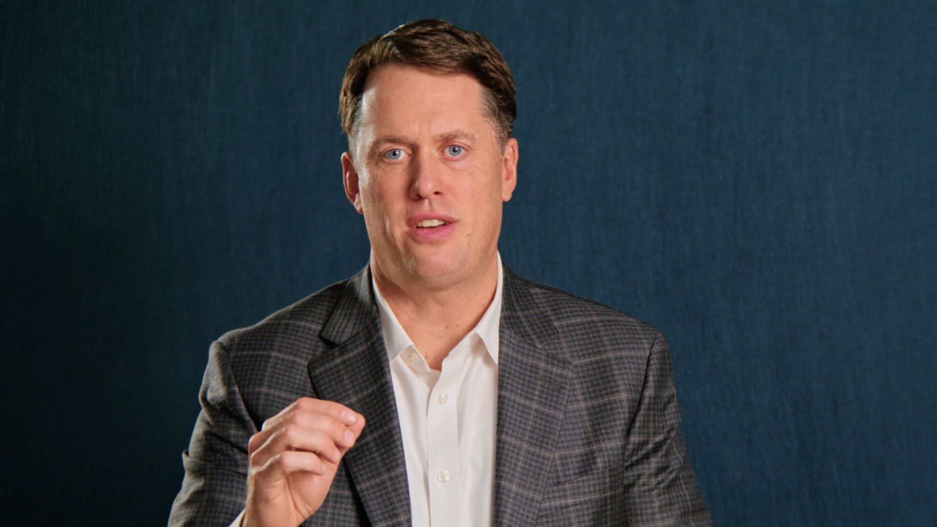 Man in gray blazer and white shirt speaks against a blue background, gesturing with his hand.