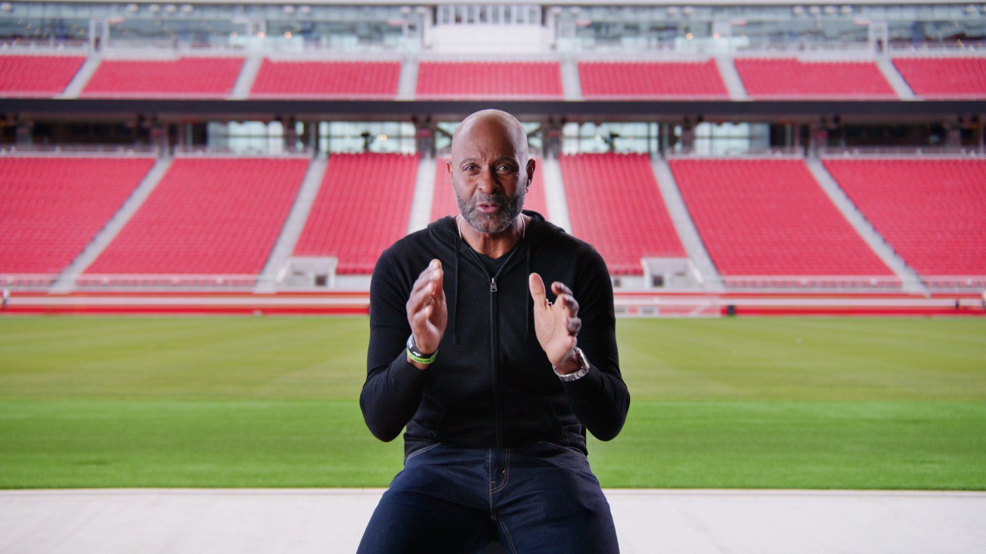 Man seated, gesturing with hands, in front of a stadium background. He's wearing black, looking forward.