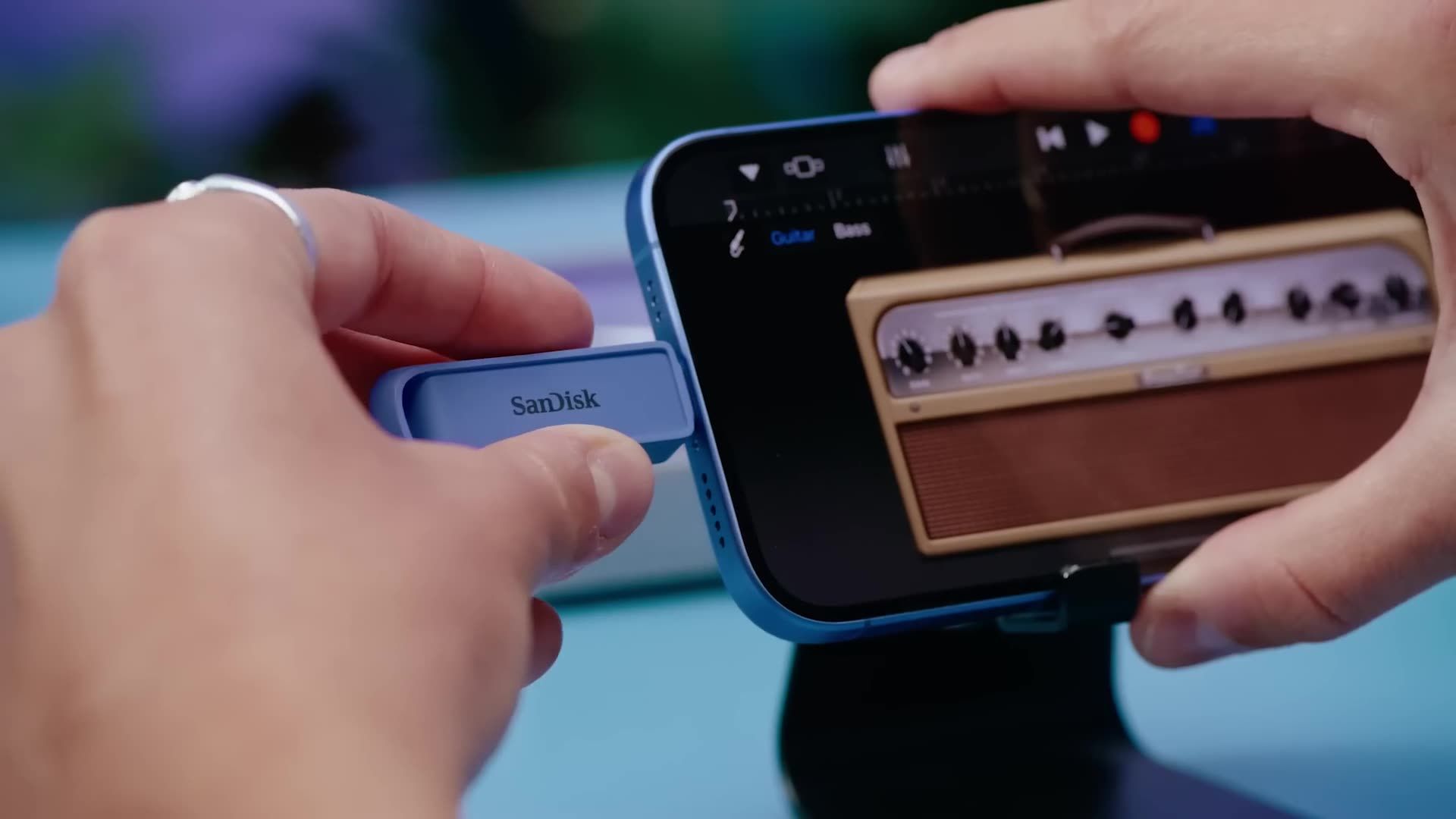 Hands holding a blue phone, connected to a blue device. The phone displays an image of an amplifier.