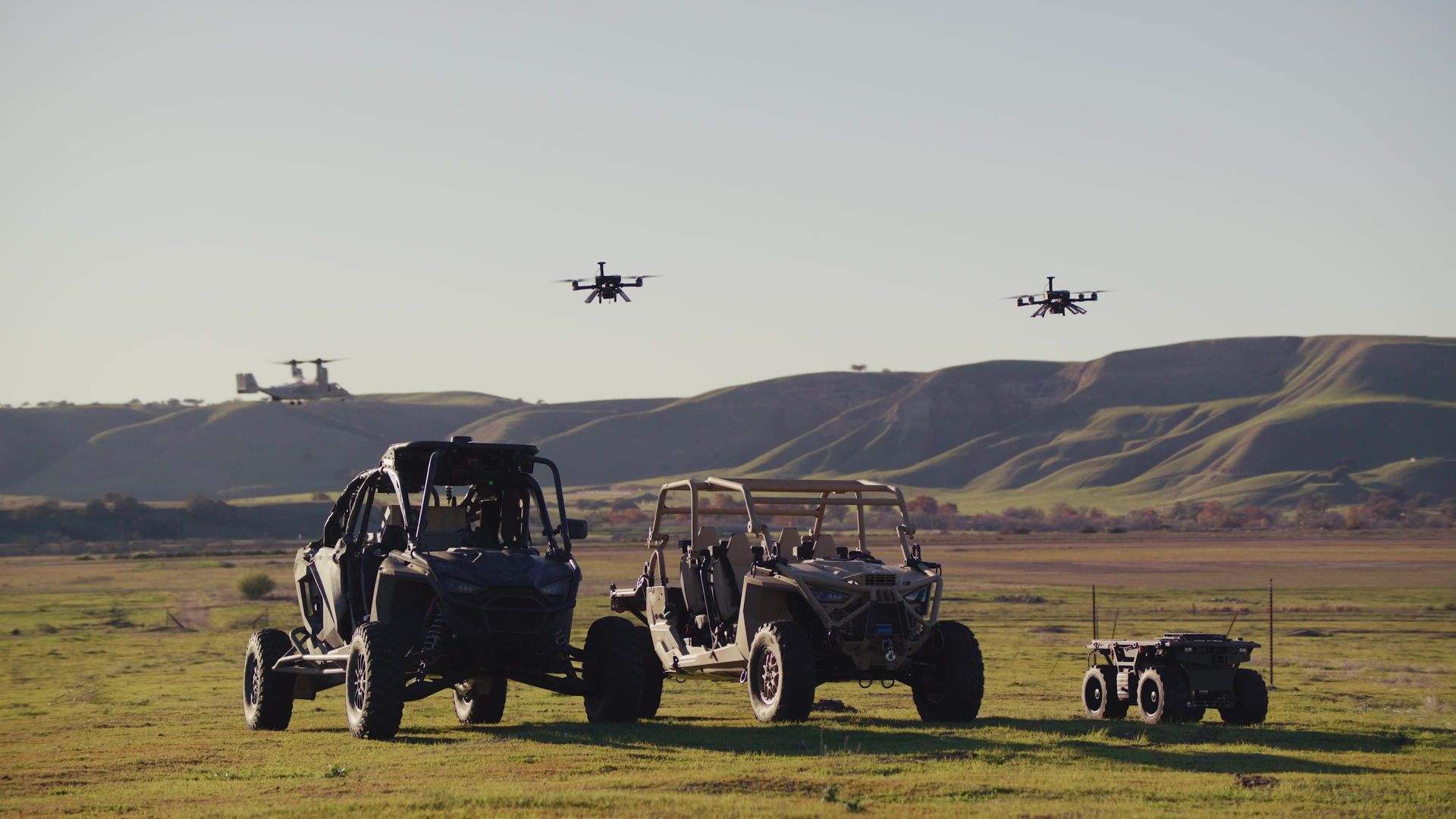 Two drones fly above vehicles in a field, with hills in the background.