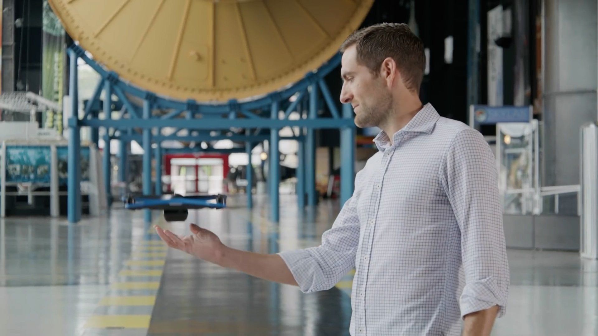 Man with outstretched hand, small drone hovering above it inside a museum with a large object in the background.