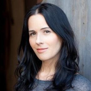 Woman with dark hair smiles, wearing a grey shirt, against a wooden background.