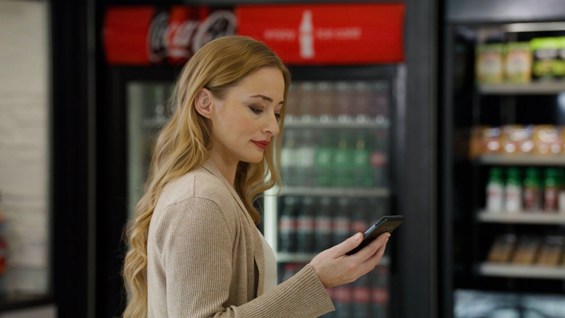 Woman looking at phone in front of a Coca-Cola cooler and store shelves.