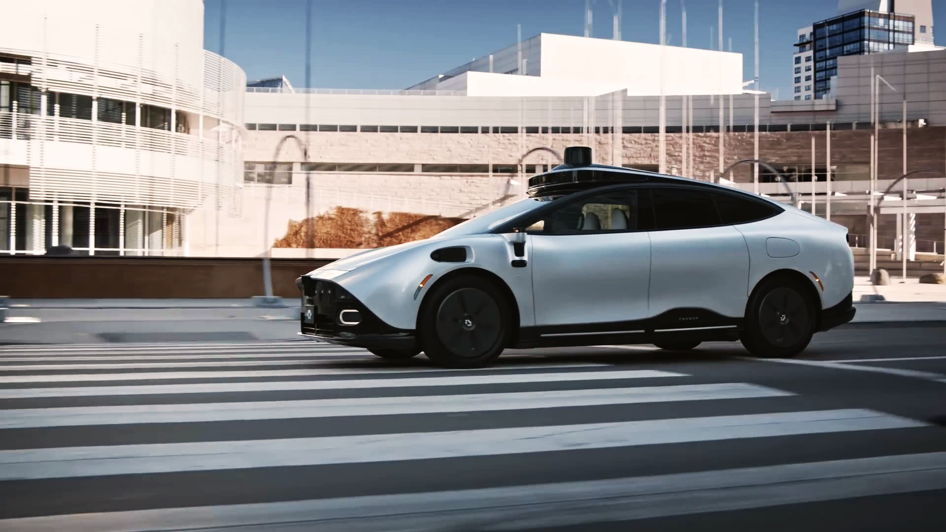 White autonomous car driving across a crosswalk in an urban setting.