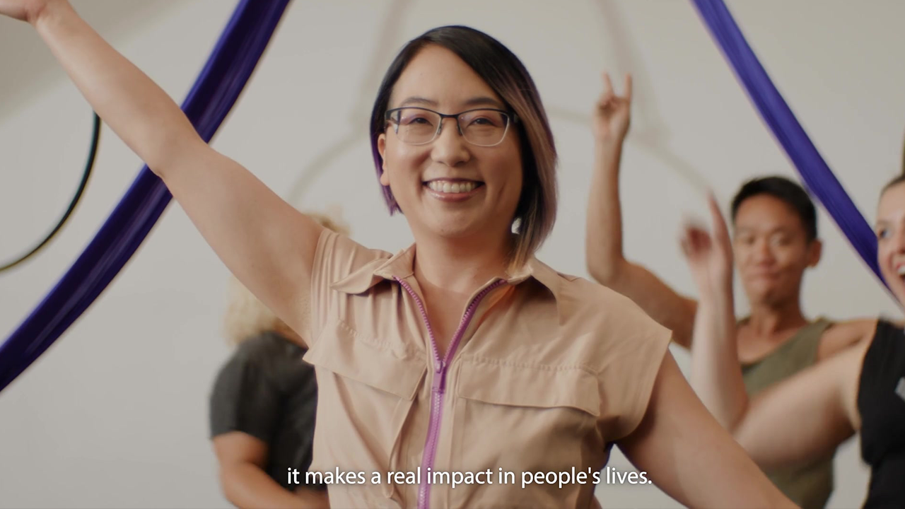 Woman in glasses smiles, arms raised, with a group in a studio with purple fabric.