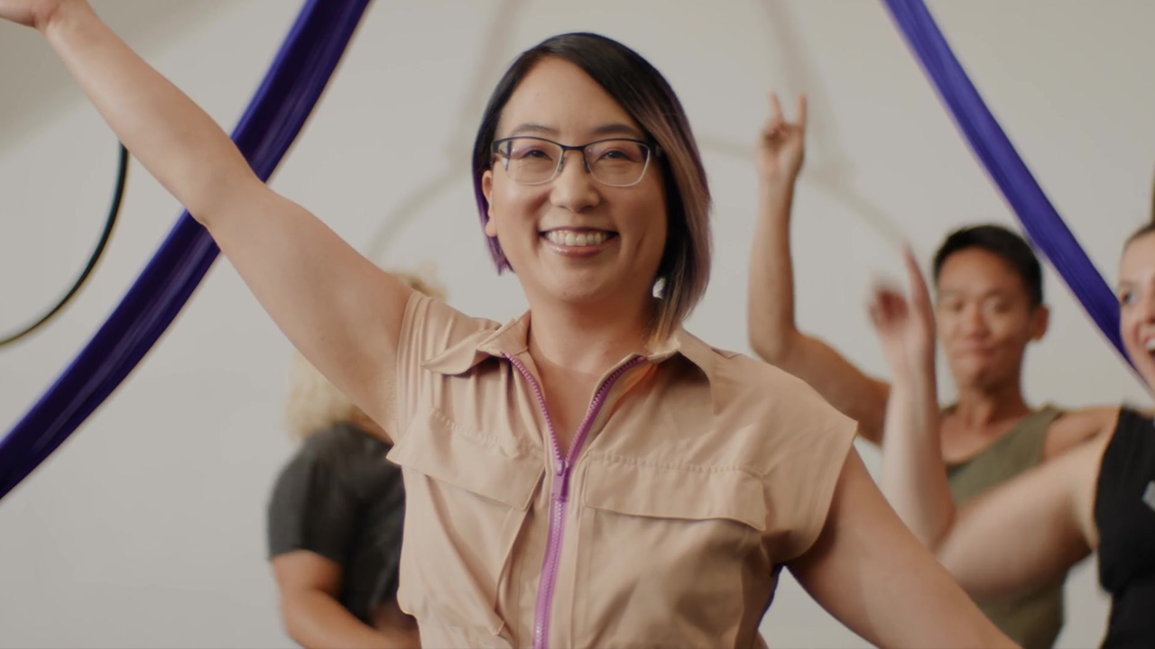 Woman with arms raised, smiling, in front of others in a dance studio. Purple aerial silks are visible.