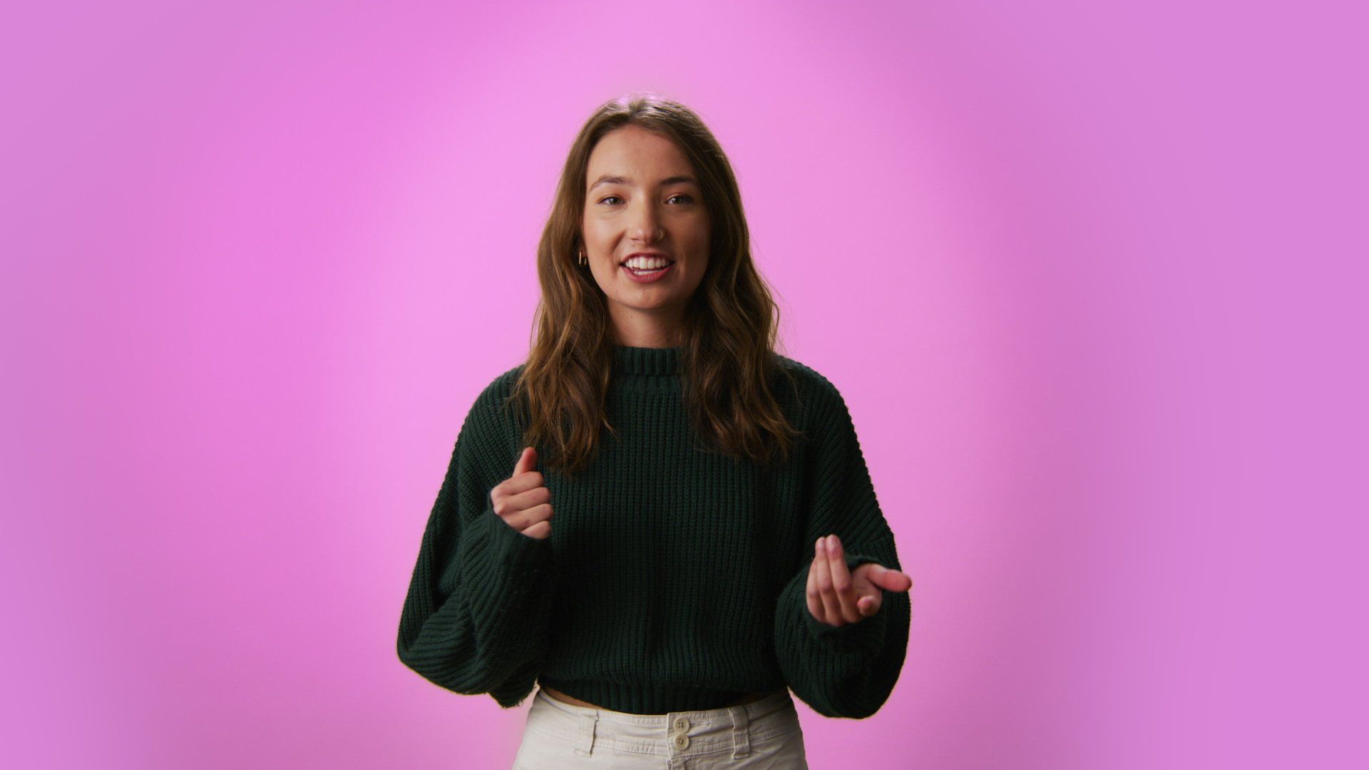 Woman in green sweater gestures with hands against a pink background.