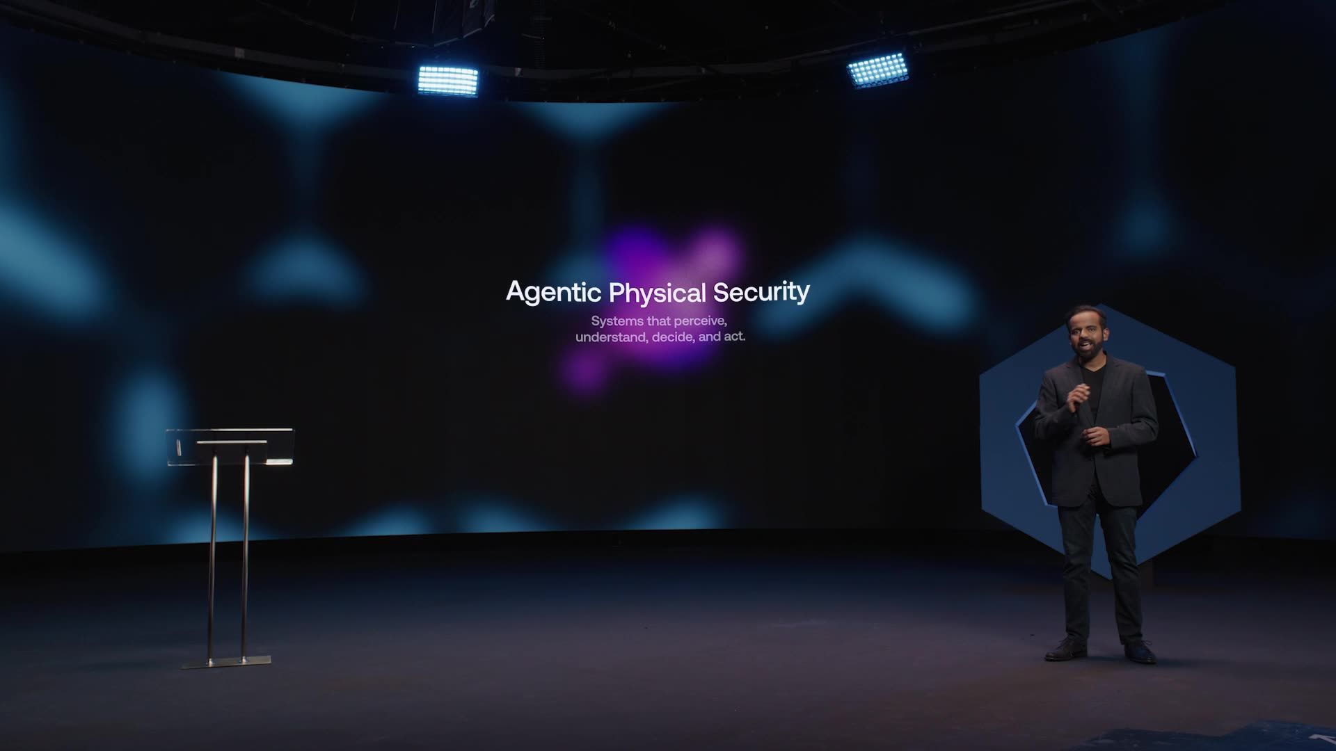 Man on stage, speaking at conference. Title: Agentic Physical Security. Blue and purple background.