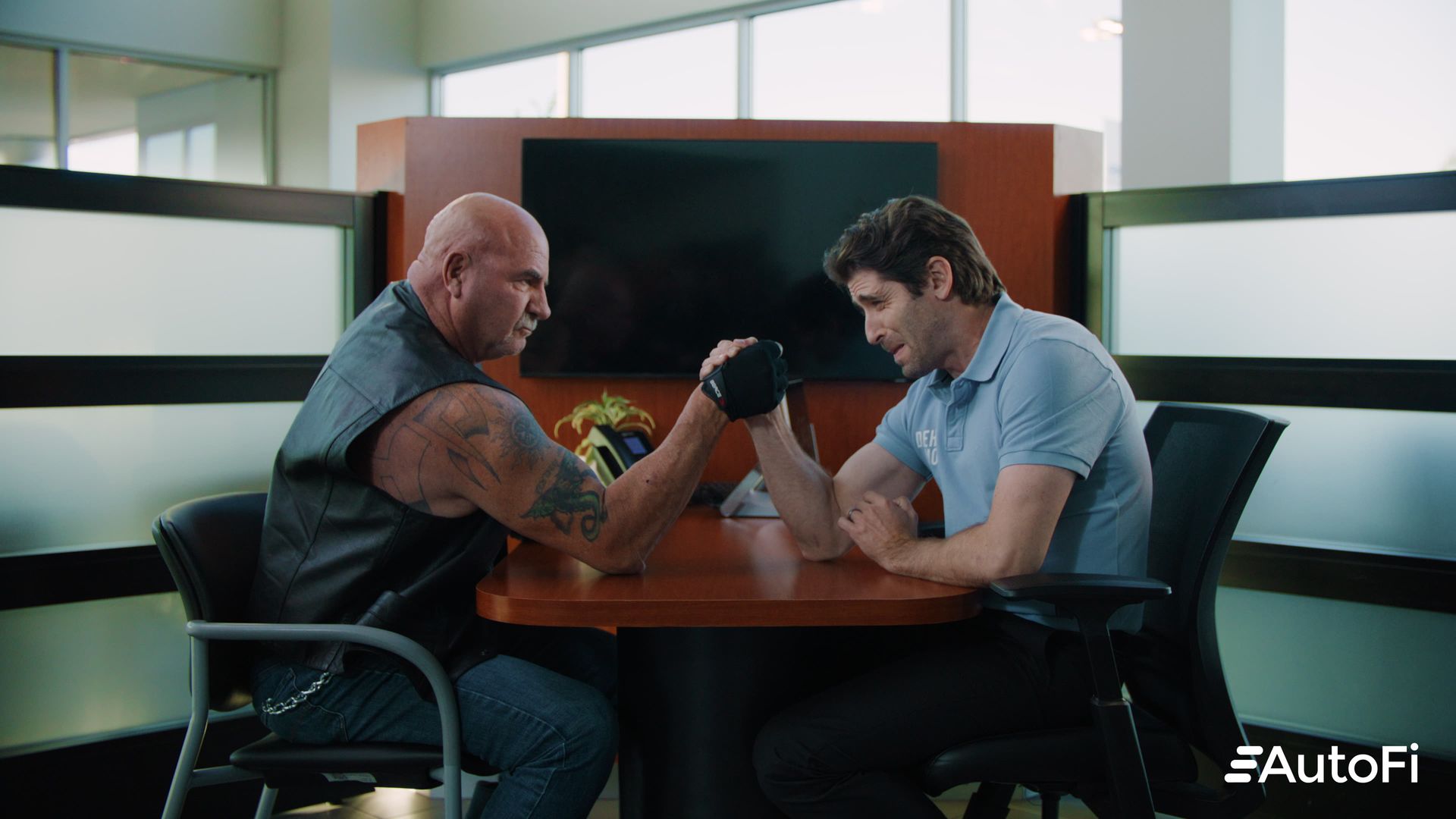 Man and muscular man arm wrestle at a table in an office setting.