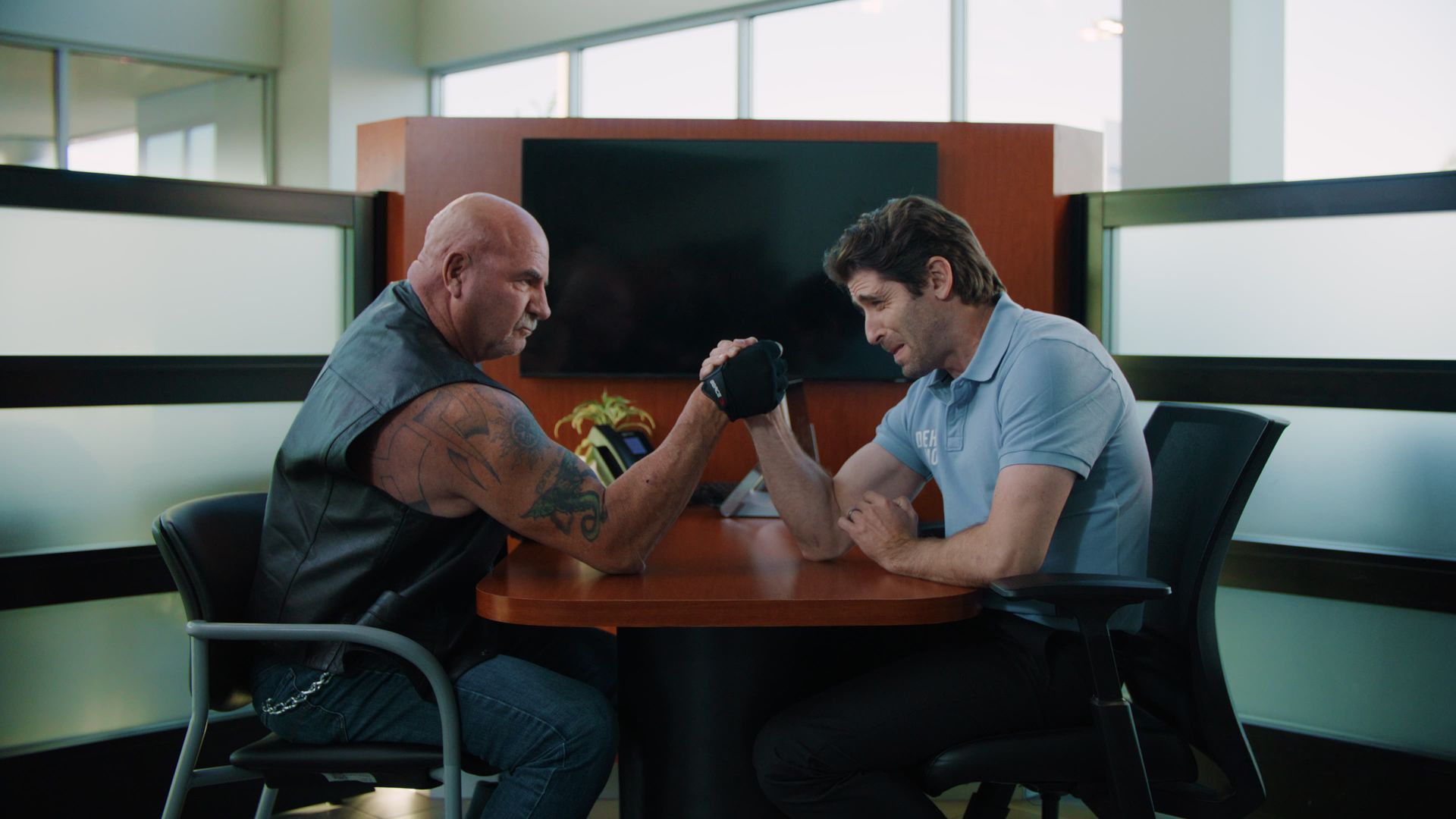 Man and muscular man arm wrestle at a table in an office setting.