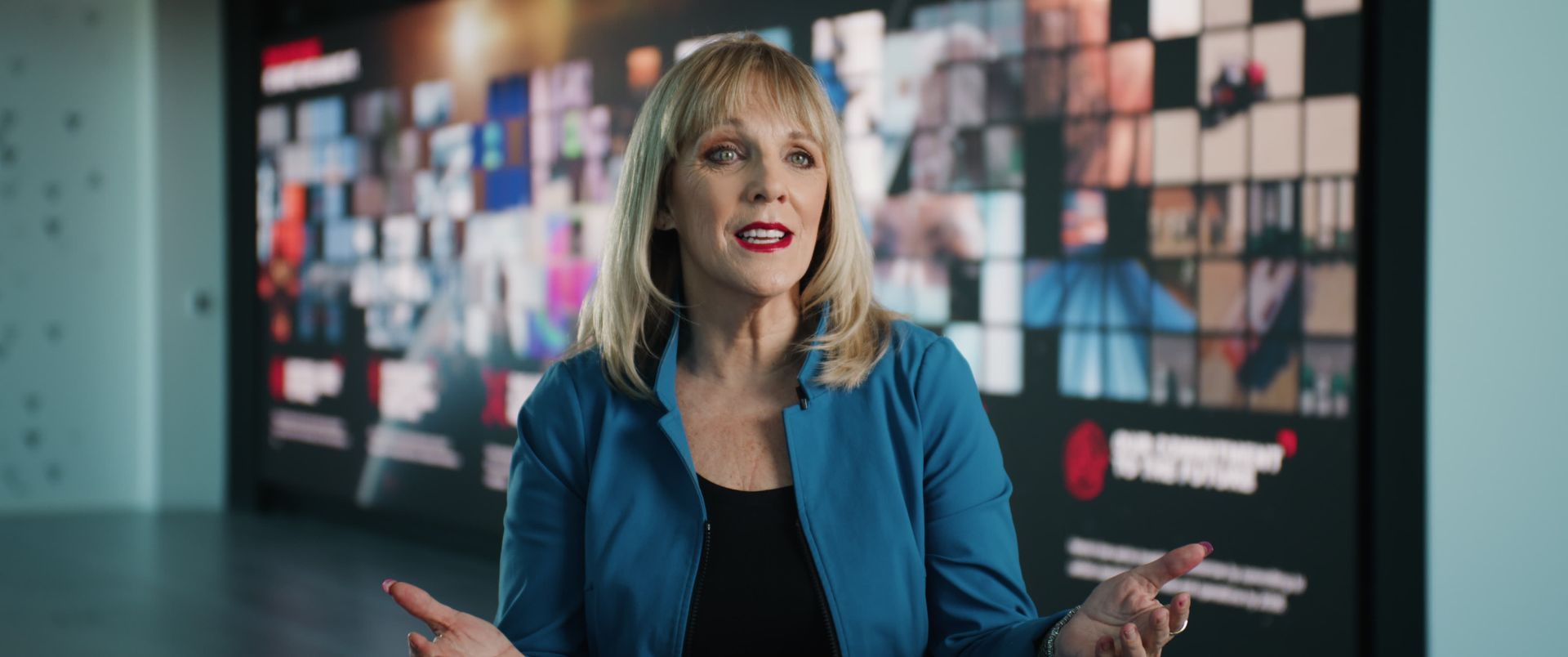 Woman in blue jacket gesturing in front of a wall of screens.
