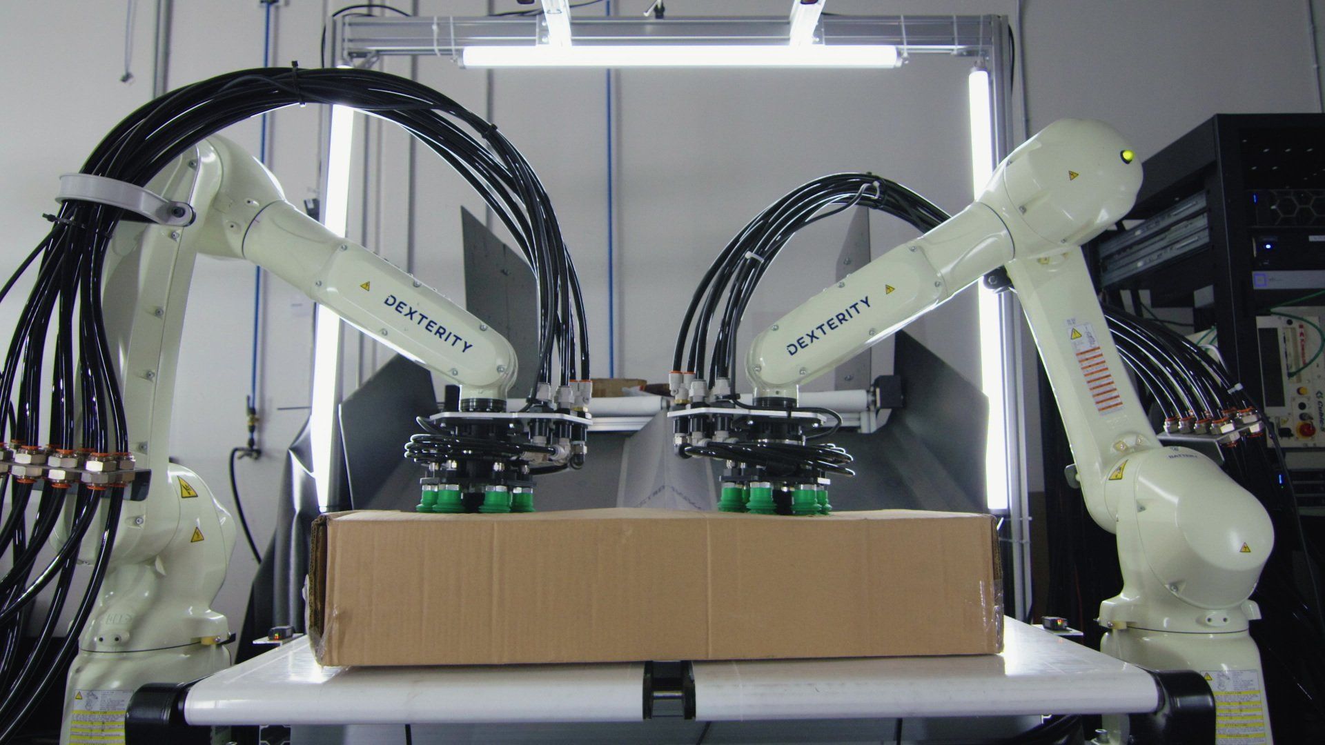 Two white robotic arms grasping a cardboard box on a white table in a lab setting.