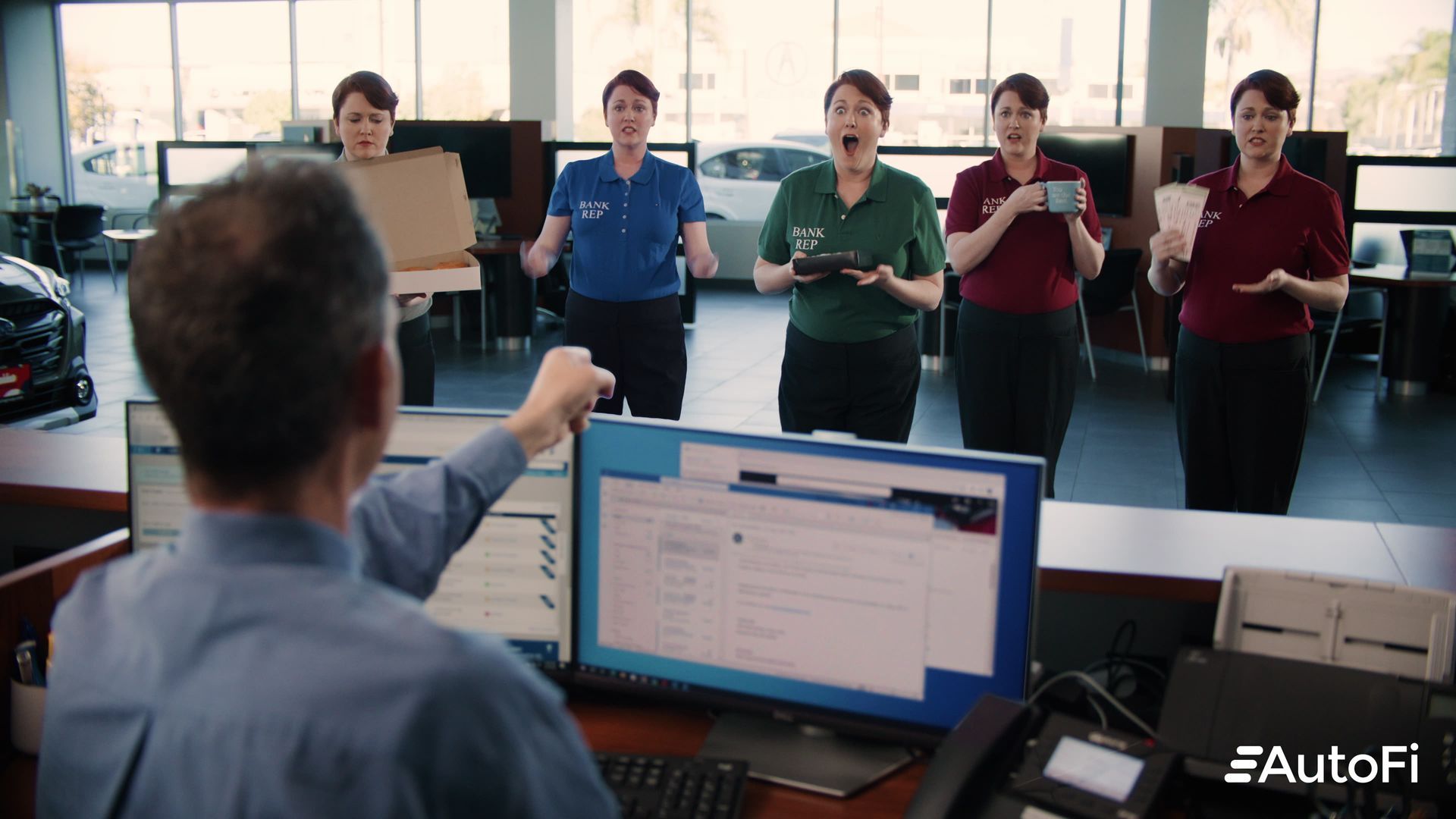 Man at desk points to a computer screen; five women react excitedly in an office setting.