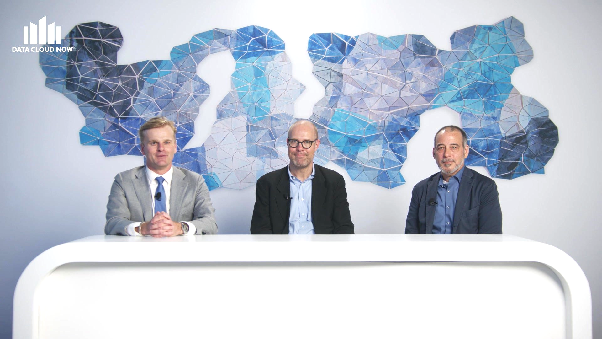 Three men seated at a white table in front of a blue and white geometric design; all wearing suits.