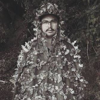 Man in camouflage suit and hat stands in front of a tree.
