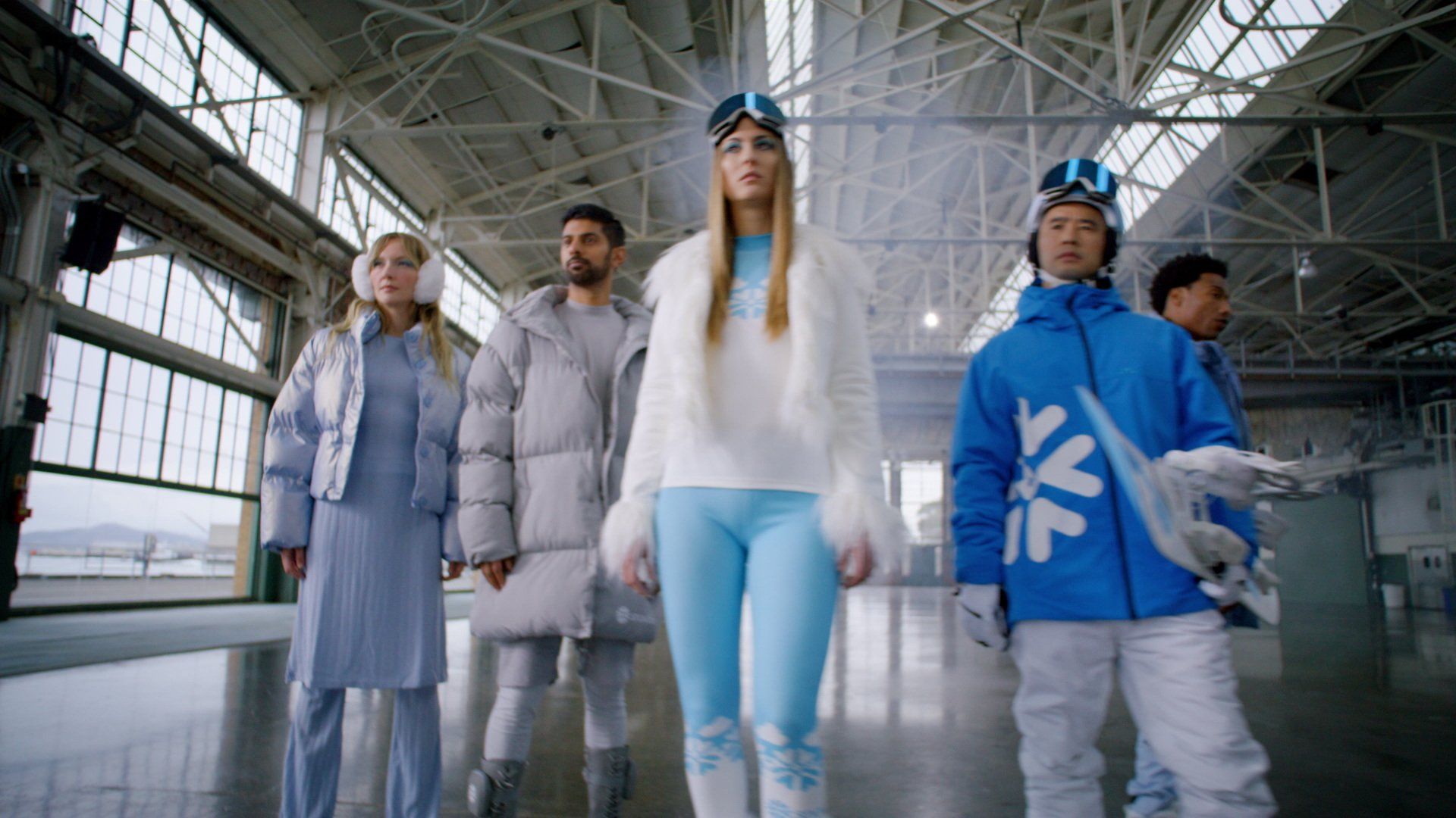 People in winter apparel stand inside a large industrial building, wearing various shades of blue and white, and looking forward.