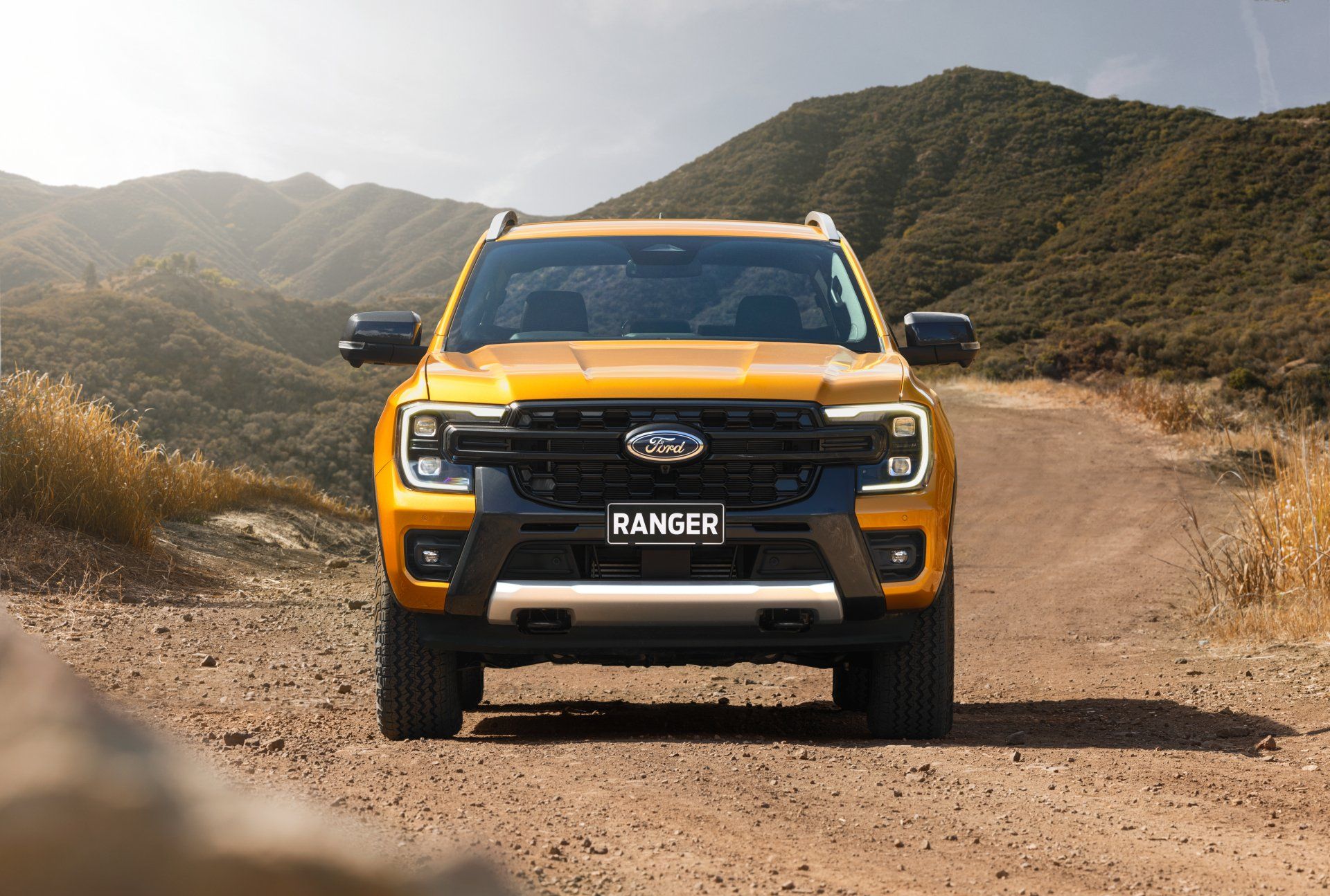 Yellow Ford pickup truck on a dirt road in a mountainous setting, facing the viewer.