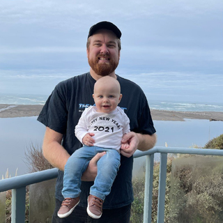Man holding a baby, both smiling, with a coastal landscape in the background. Baby wears a