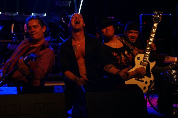 Vier muzikanten op het podium onder blauwe en paarse lichten, een van hen speelt gitaar, de anderen glimlachen en zingen.