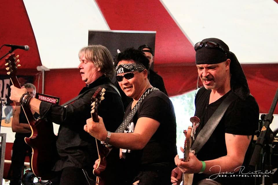 Drie muzikanten treden op het podium op, twee bespelen een gitaar en één zingt in een microfoon.