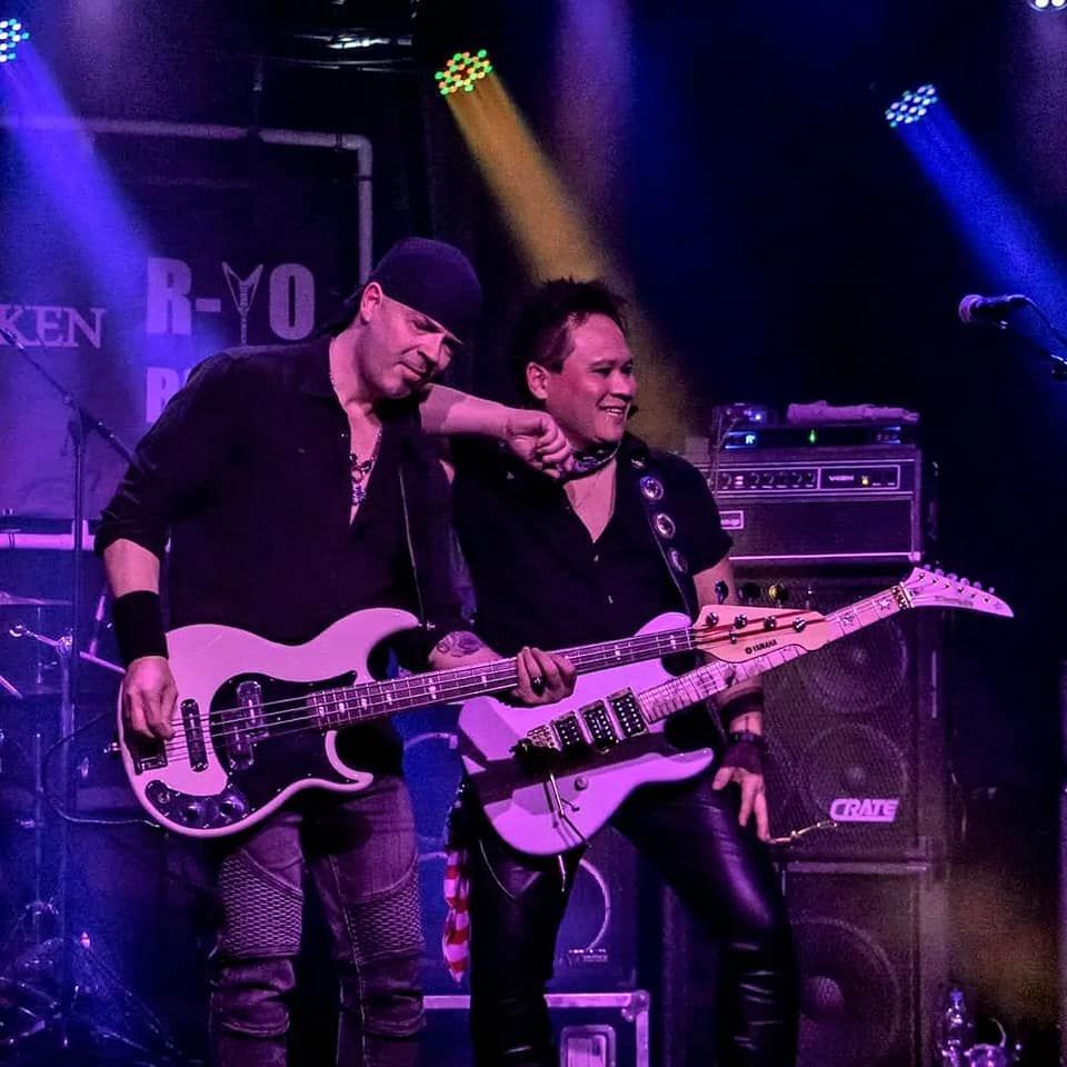 Twee gitaristen treden op het podium op onder paarse en blauwe lichten, lachend naast elkaar.