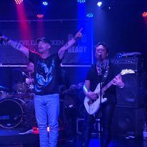 Twee muzikanten op een blauw verlicht podium, de ene wijst omhoog en de andere speelt op een witte gitaar.