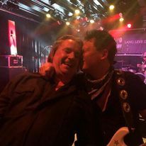 Twee mensen glimlachen en lachen samen op een concertpodium met kleurrijke verlichting.