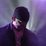 Muzikant met zwarte pet en jasje onder paarse podiumverlichting, naar beneden kijkend.